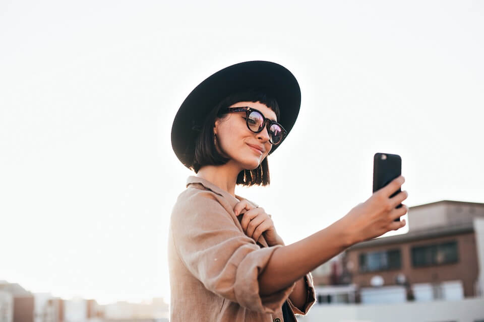 Studentin der ECBM European College of Business and Management macht ein Selfie mit ihrem Handy und traegt einen Hut und Brille im Hintergrund Gebaeude